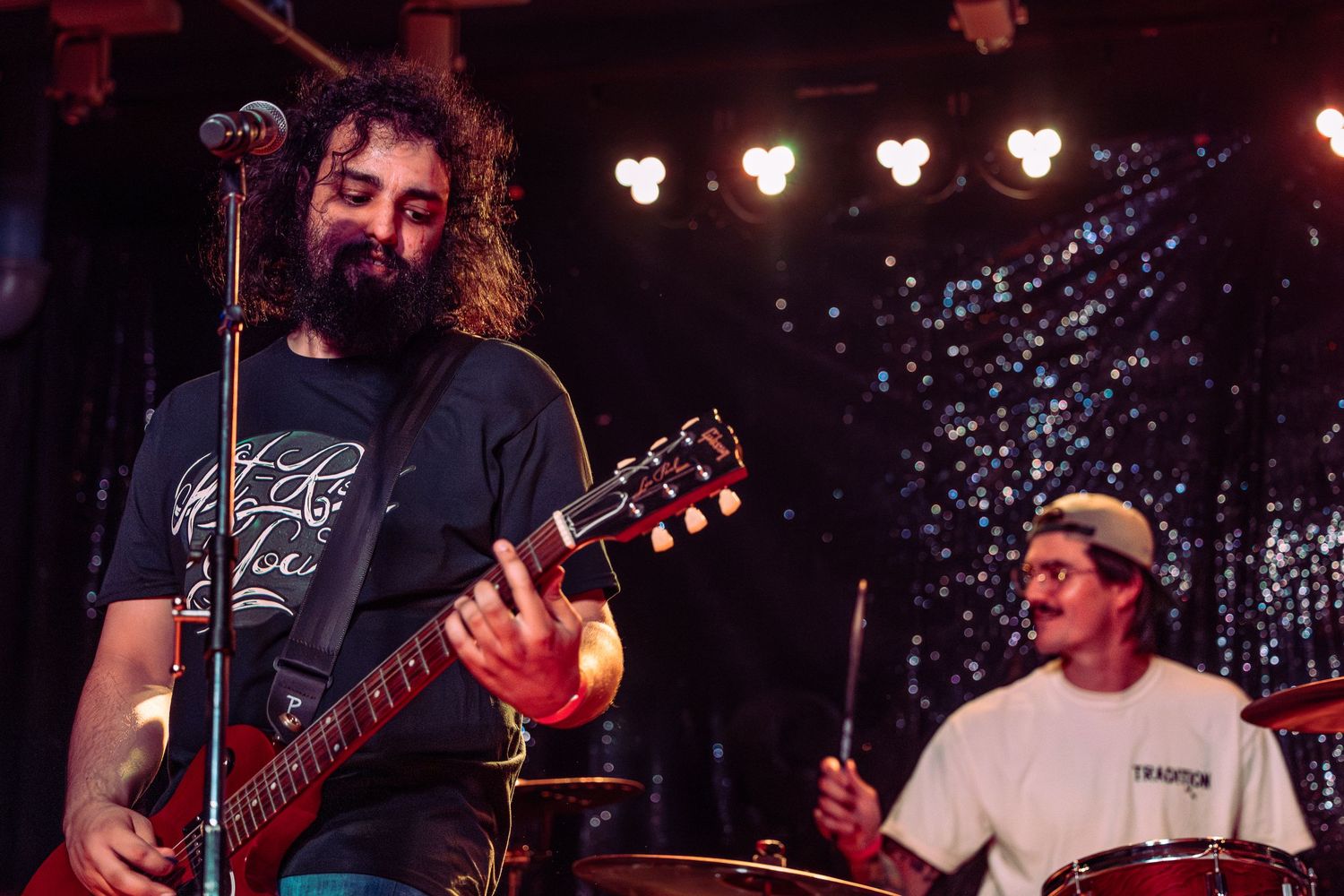 Curly-haired guitarist in dark shirt plays Gibson guitar on stage with drummer visible in background.