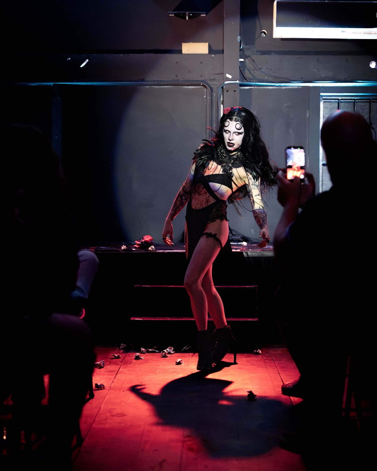 Tattooed gothic performer dances under red stage lighting wearing black lingerie and feathered costume.