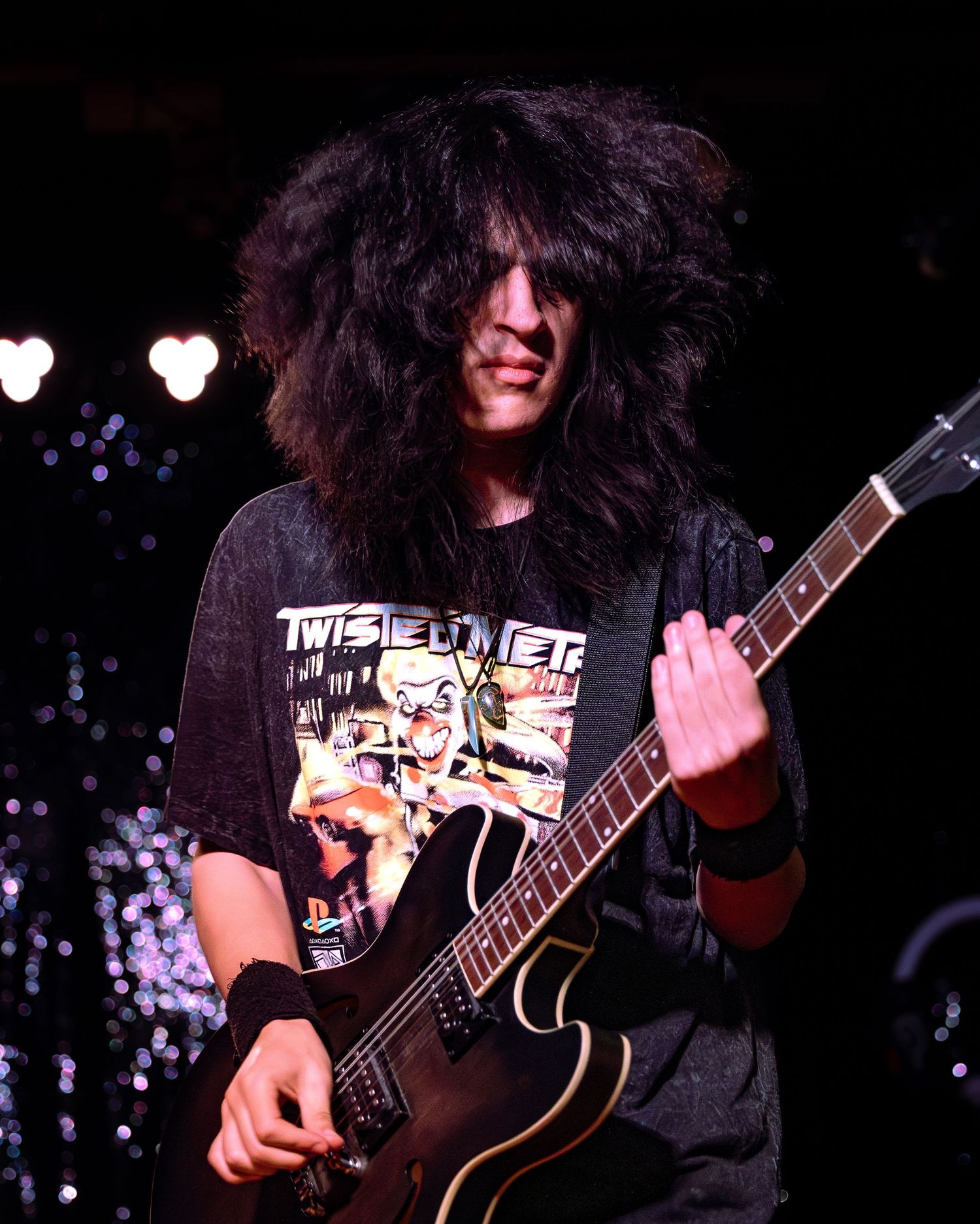 Long dark-haired guitarist playing a black electric guitar on a dimly lit stage.