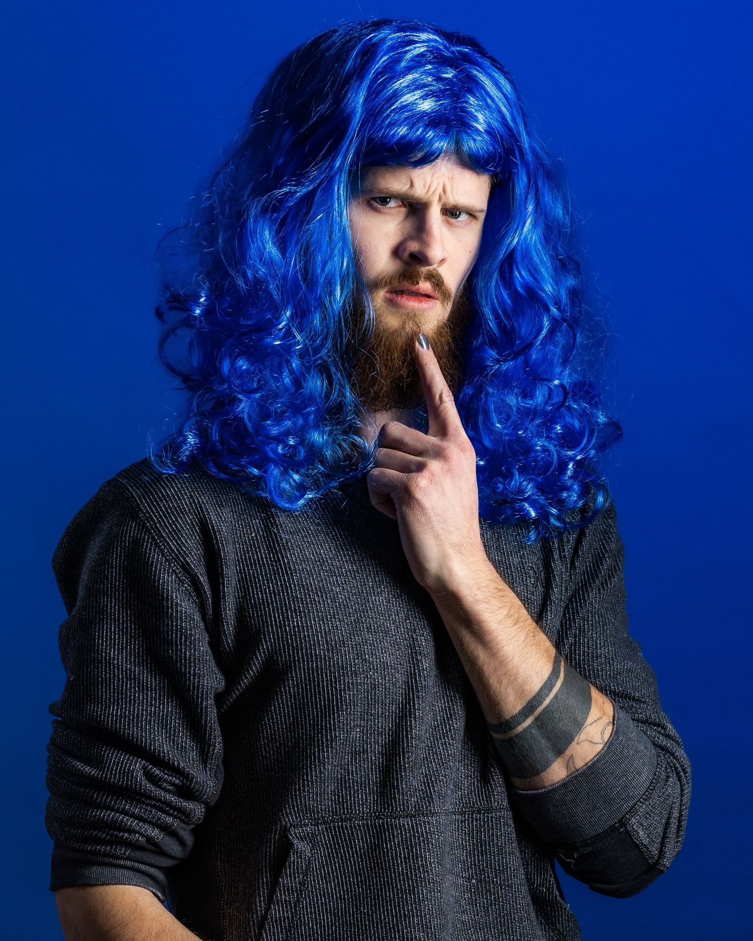 A person with bright blue curly wig and beard wearing a dark shirt poses against a vibrant blue background.