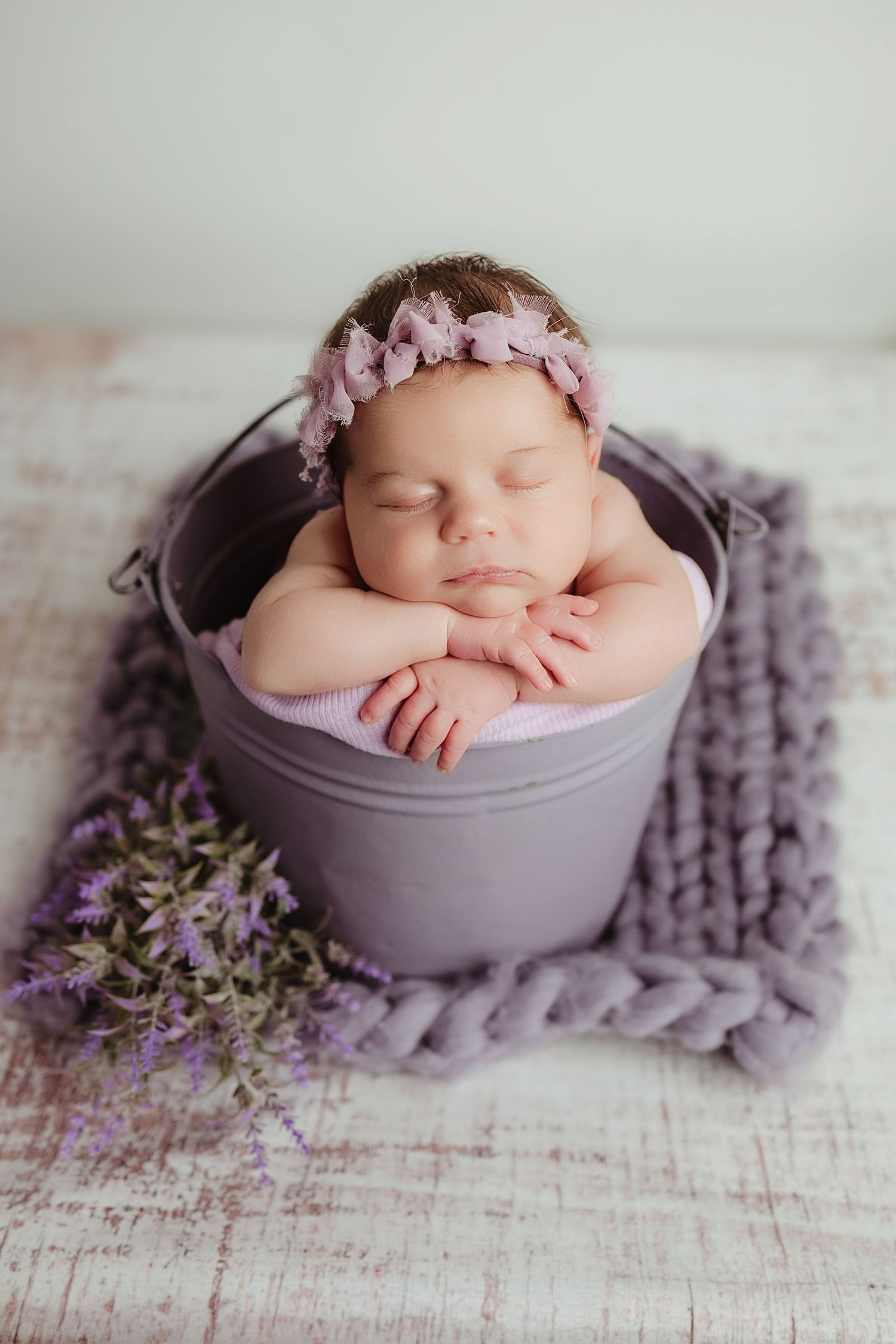 Sleeping newborn baby girl in purple bucket with floral headband on lavender knit blanket.