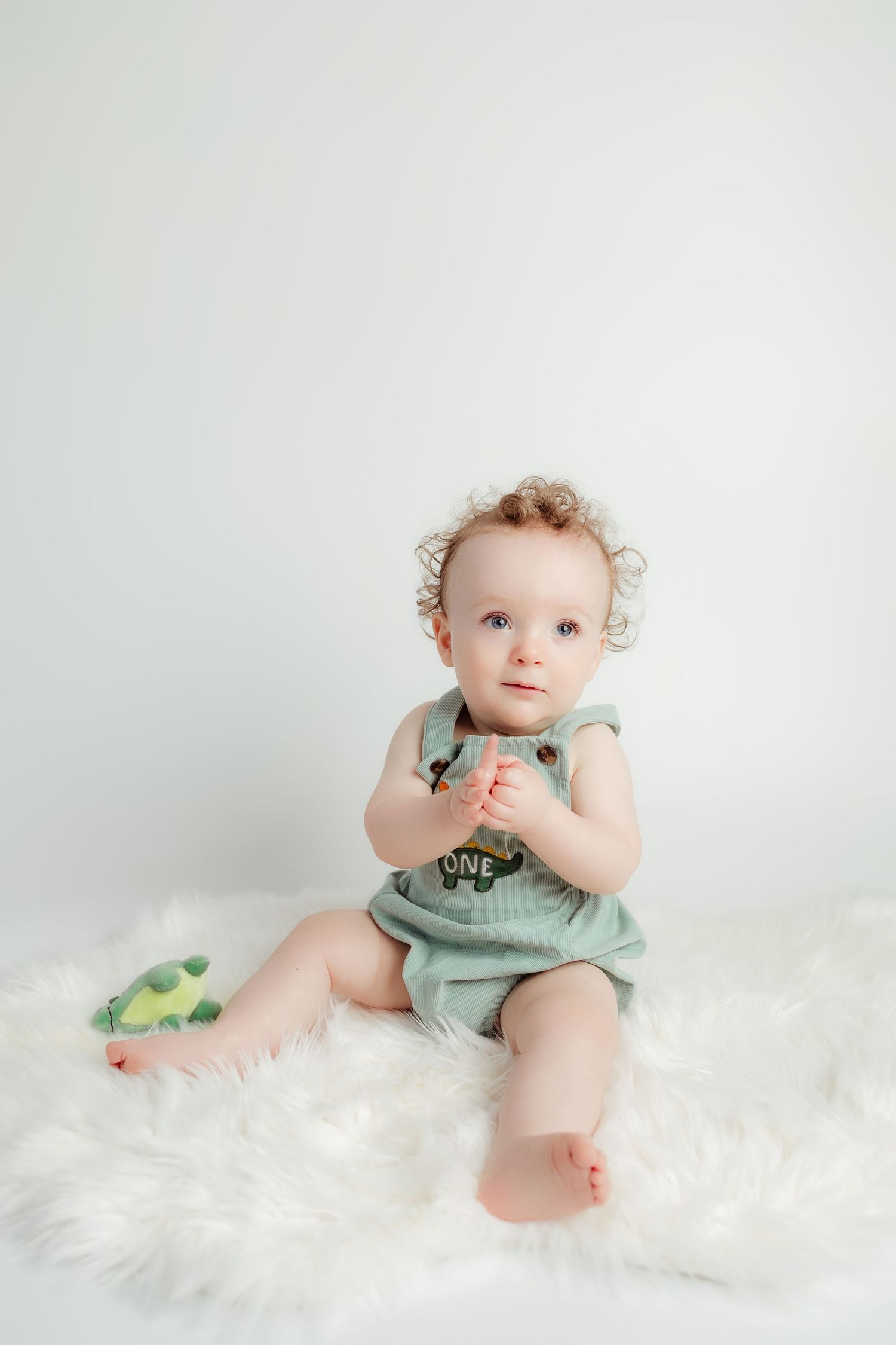 Curly haired baby in green one romper sitting on fluffy white rug with dinosaur toy.