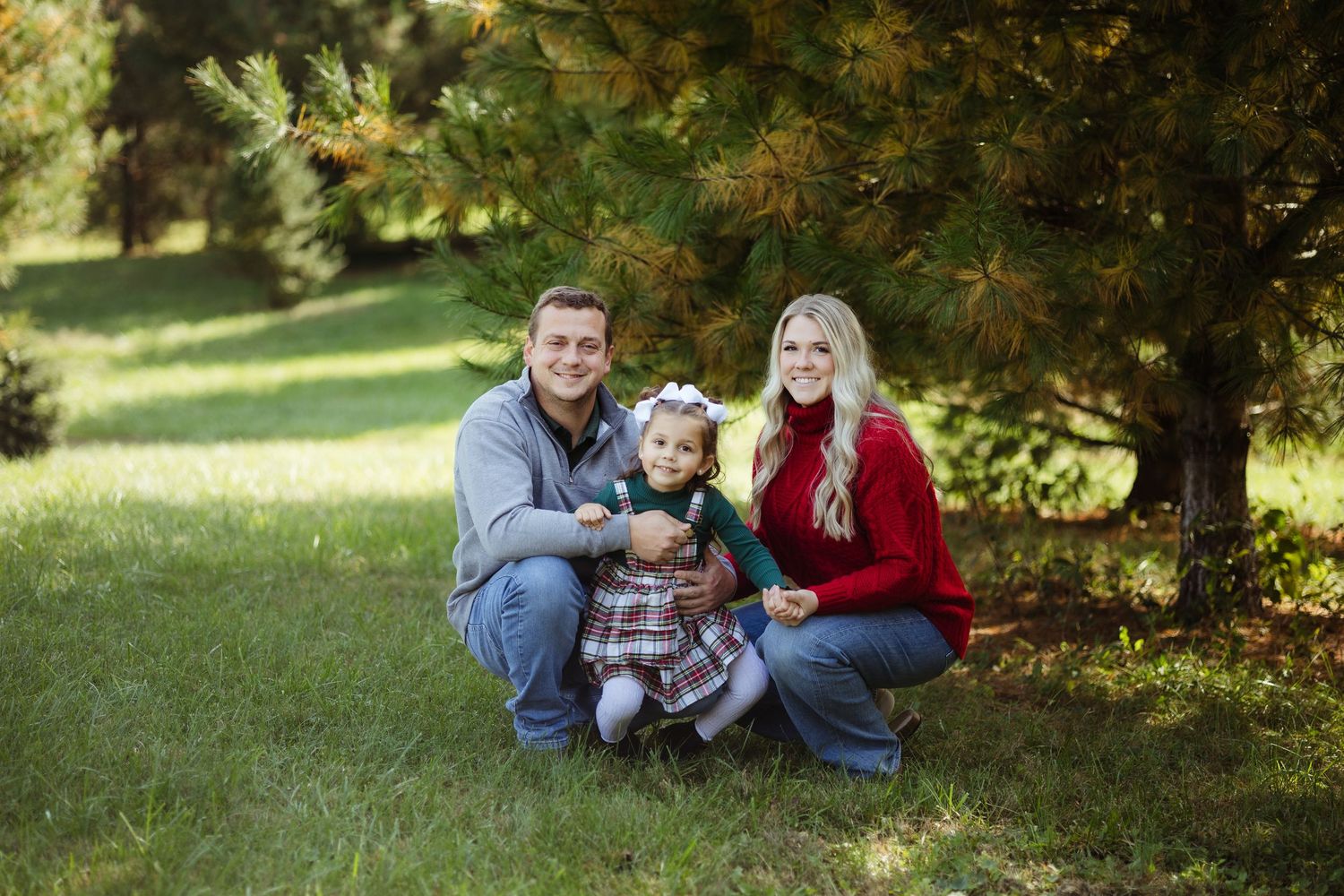 A smiling family of three crouches together on green grass surrounded by pine trees outdoors.