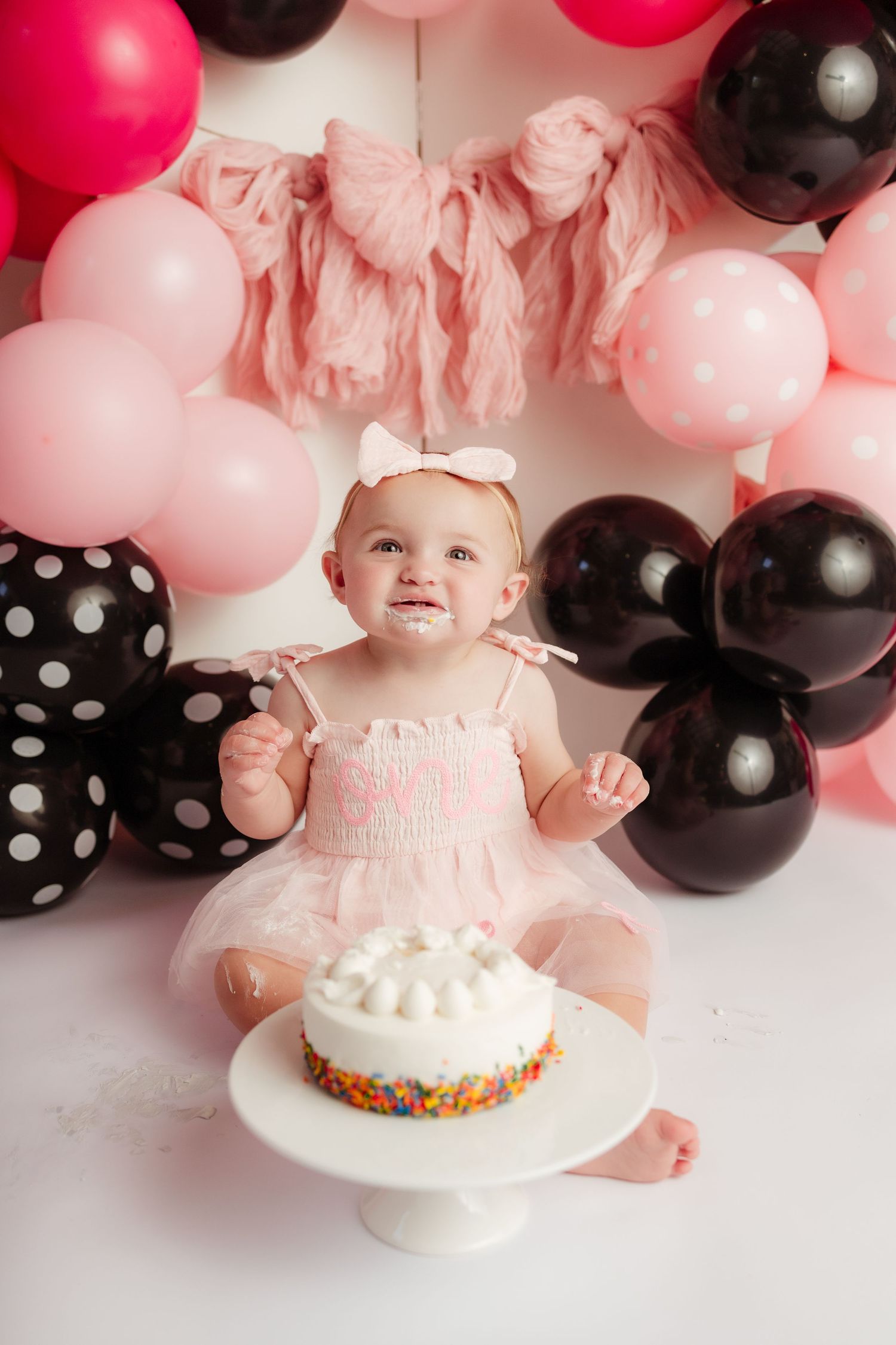 Baby girl in pink tutu dress with cake smash setup surrounded by pink and black balloons.