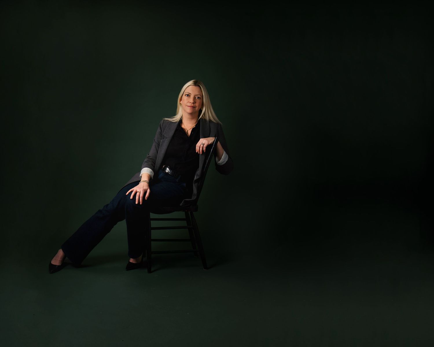 A blonde woman in dark blazer and jeans sits on a chair against a deep green background.
