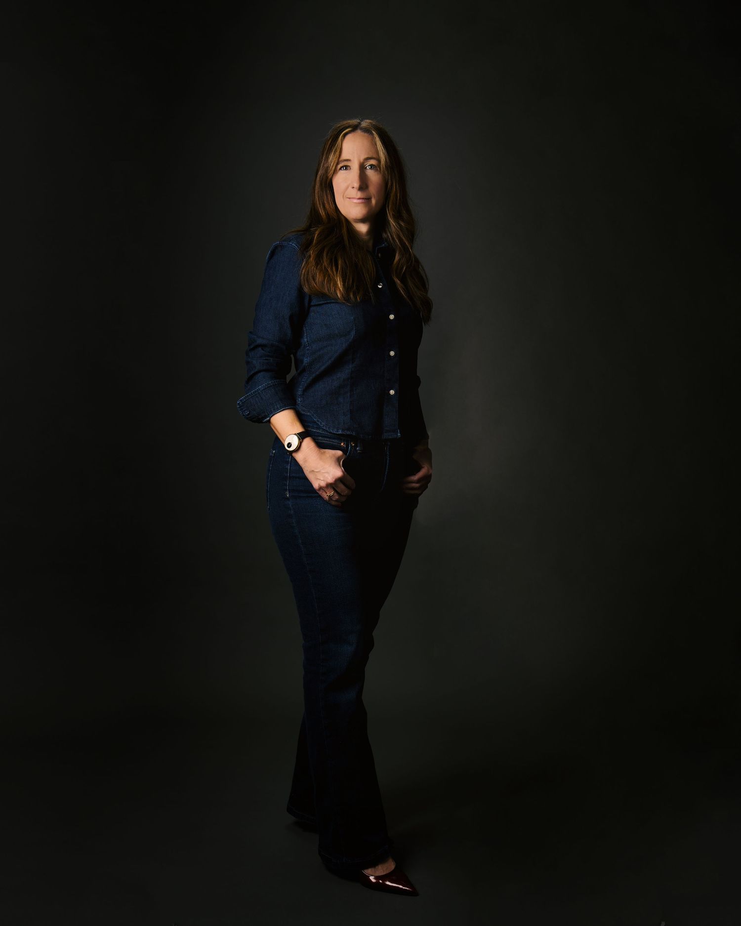 Woman in dark denim jacket and flare jeans poses confidently against dark studio background.