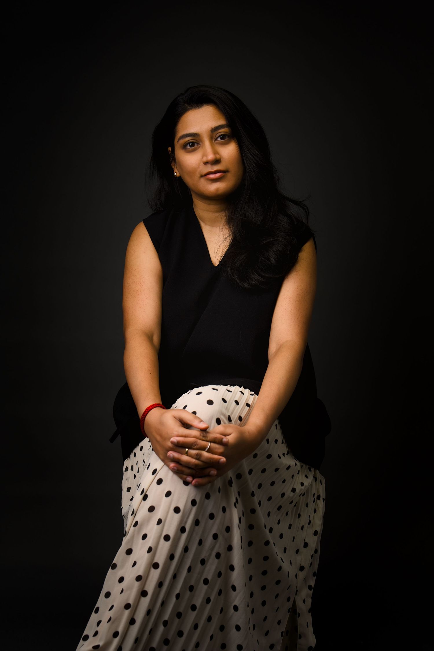 Woman in black top and polka dot skirt poses with clasped hands in dark studio.