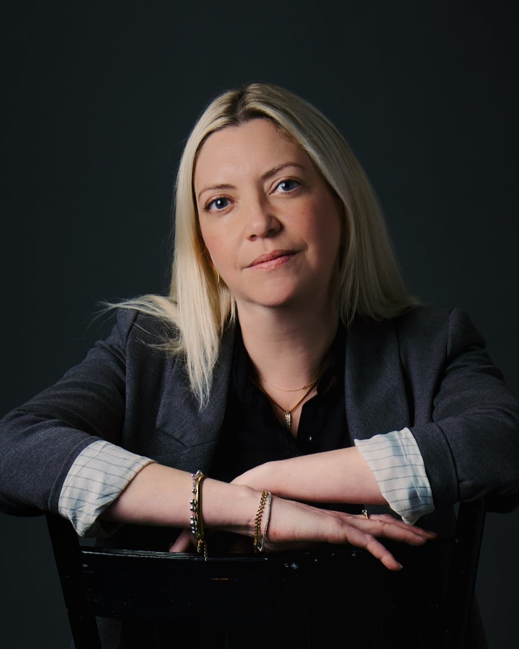 A blonde woman in a blazer leans forward with arms crossed on a chair back against a dark teal background.