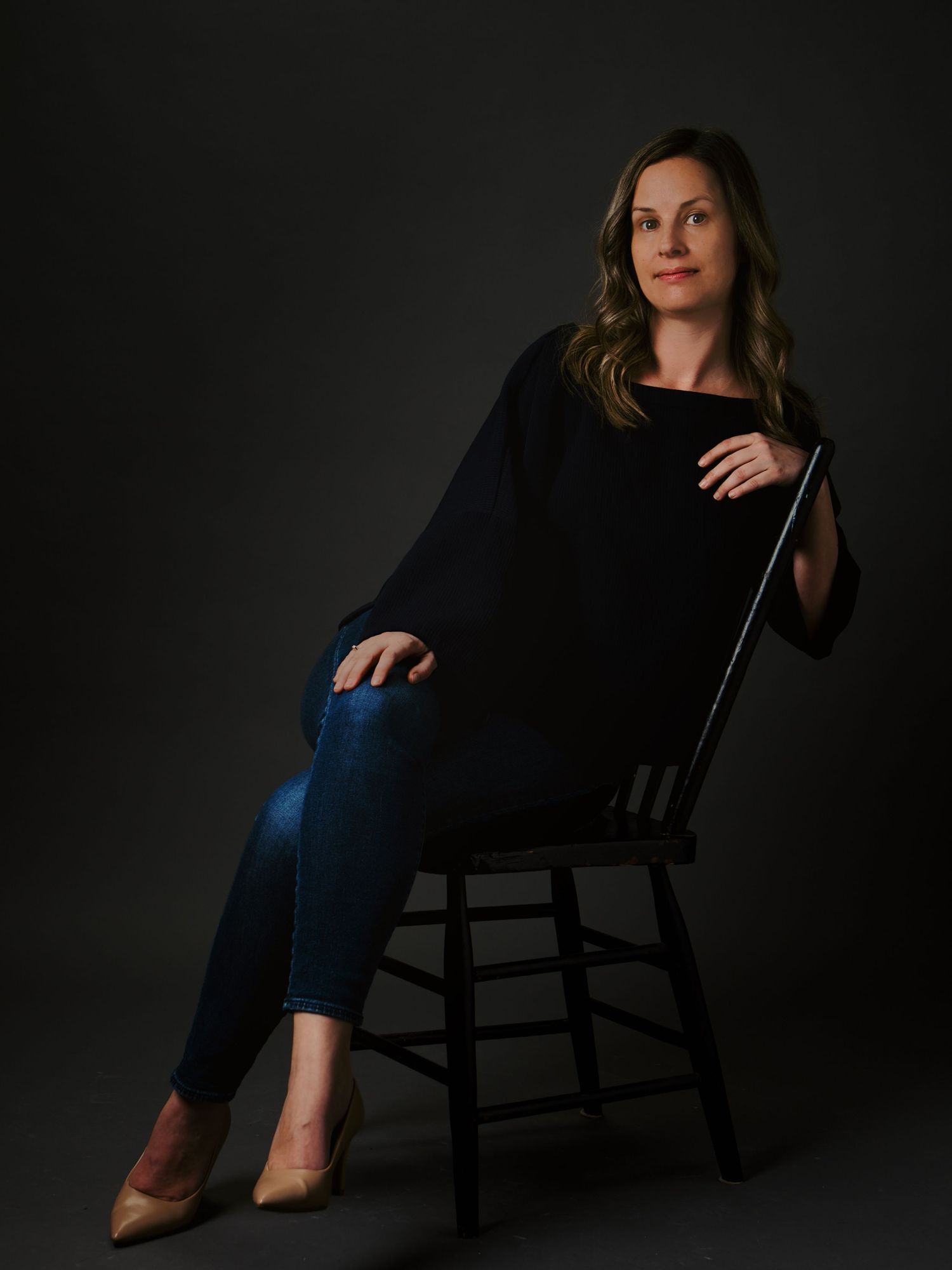 Woman in black poncho and jeans sitting on chair against dark studio background.