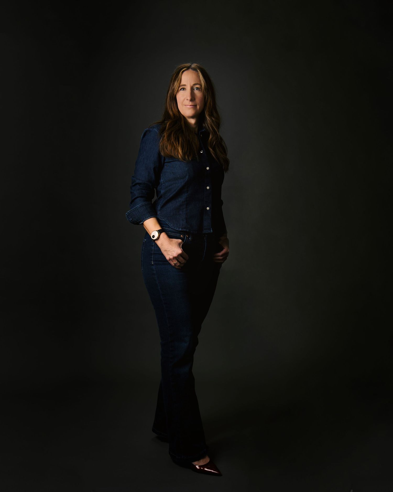 Woman in dark denim jacket and flared jeans standing against black background.