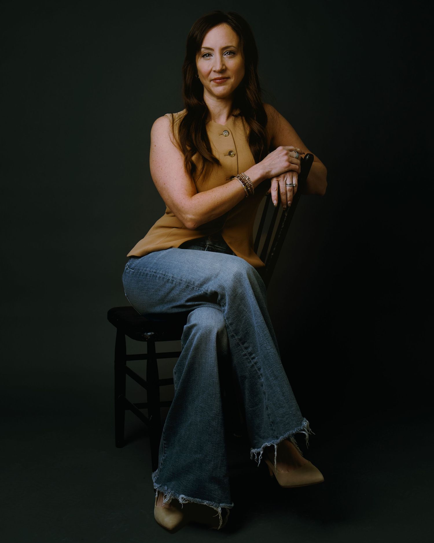 Woman in tan vest and jeans sitting on stool against dark green background.