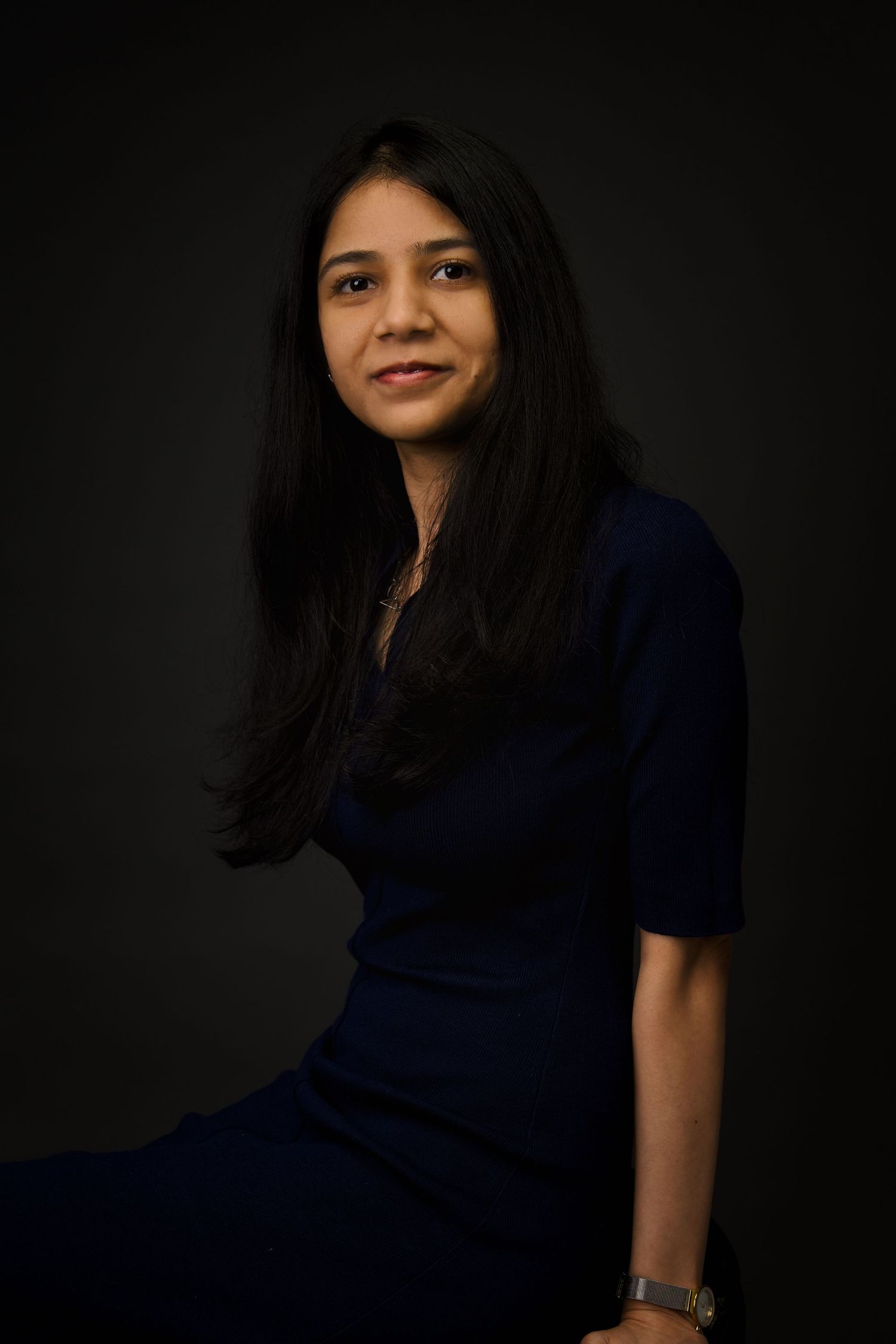 Young woman in navy blue dress poses with long dark hair against dark background.