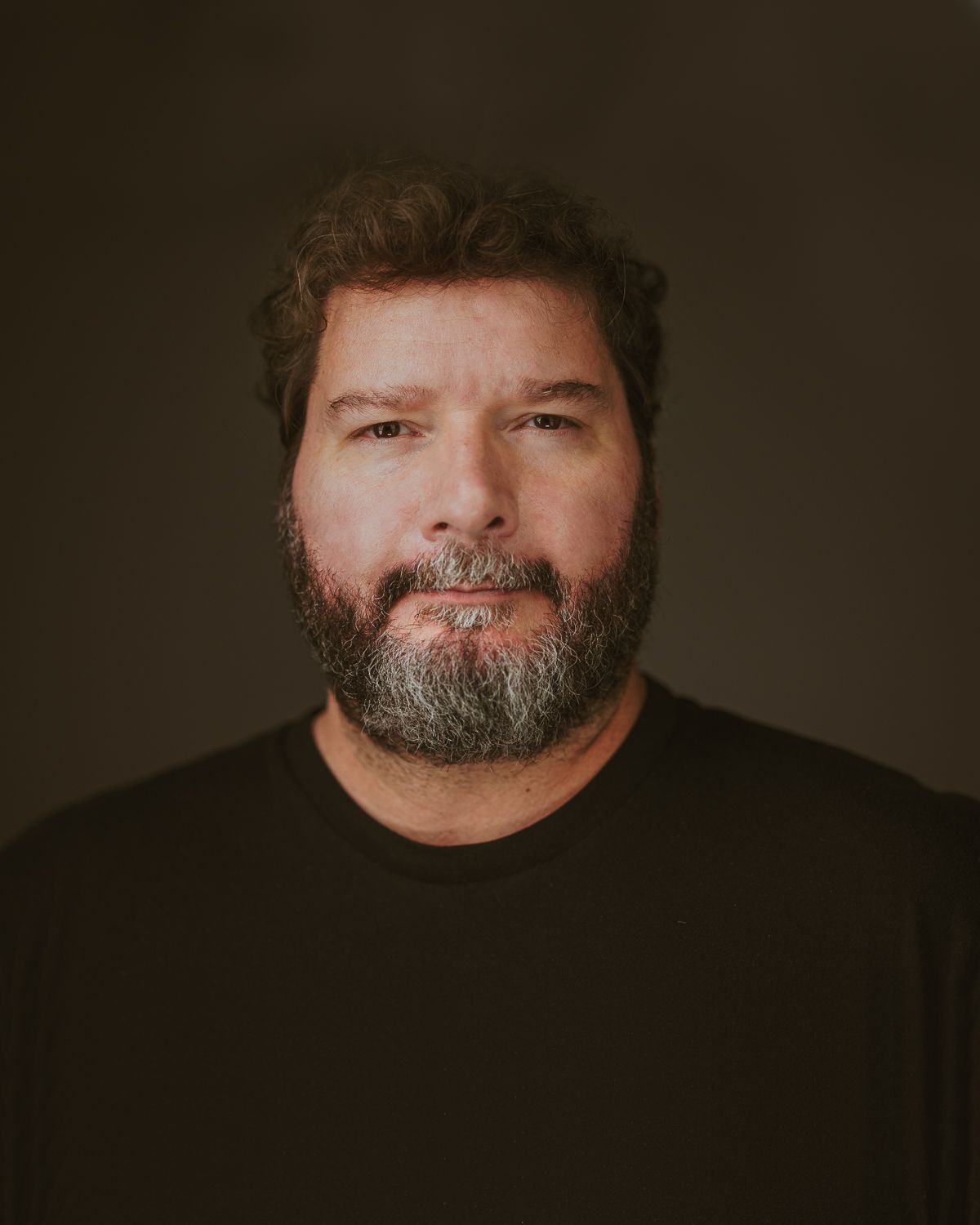 Bearded man in black t-shirt posing for professional headshot against dark background.