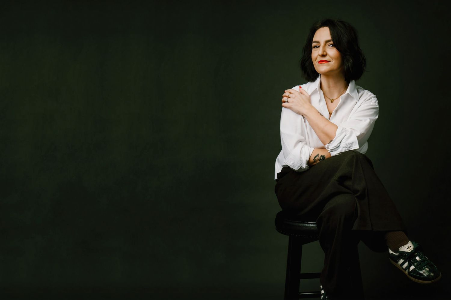 Woman in white shirt sits on stool with hand on shoulder against dark green background.
