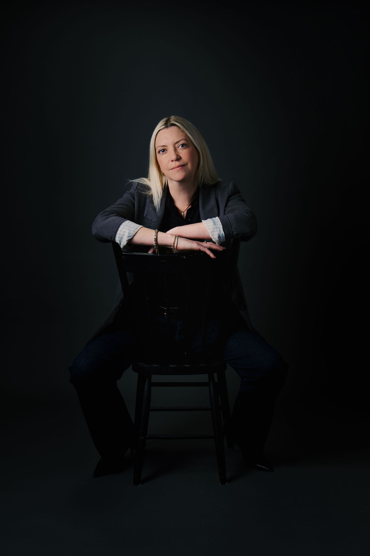 Blonde woman in gray blazer sitting on stool leaning forward against dark background.