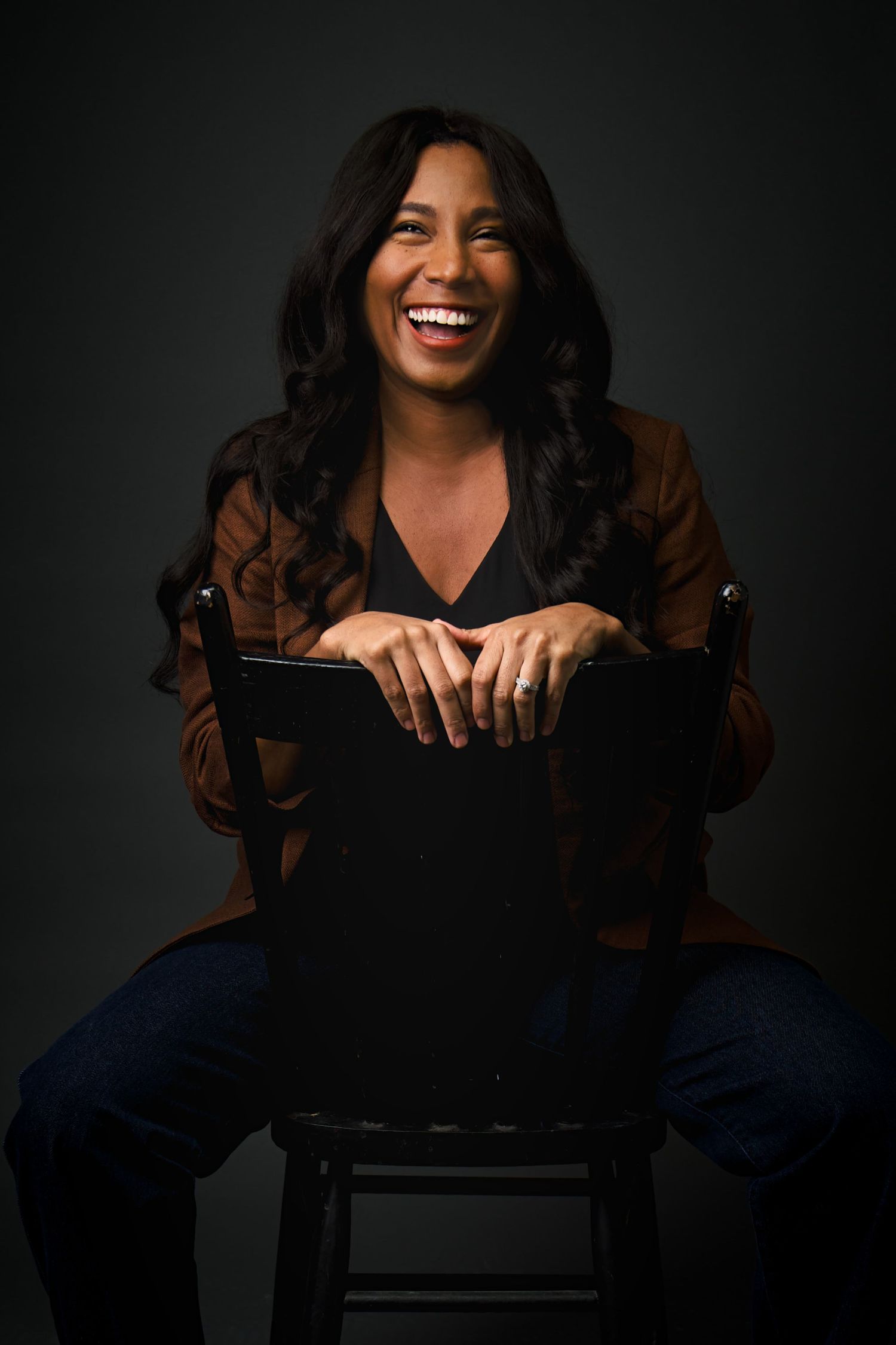 A laughing woman in a brown blazer sits confidently on a chair against a dark background.