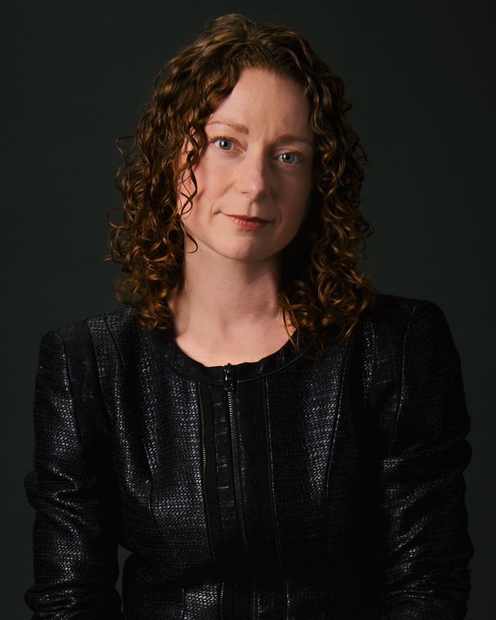 Woman with curly auburn hair in black textured blazer poses for professional portrait against dark background.