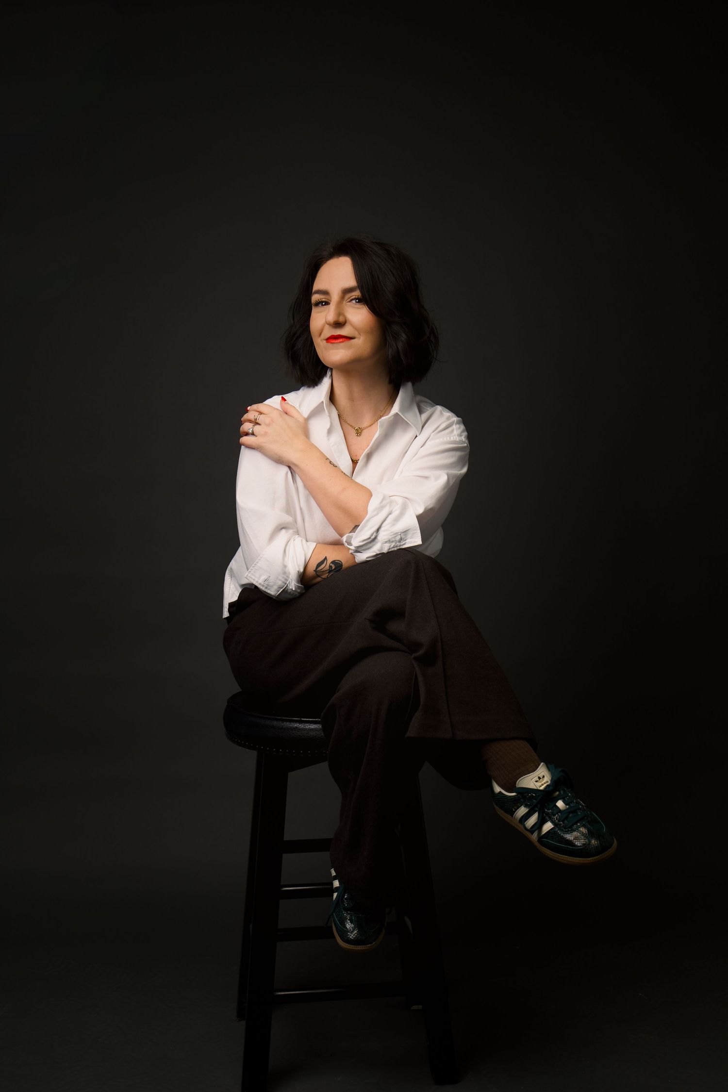 Woman in white shirt and brown pants sits on stool with hand on shoulder in studio.