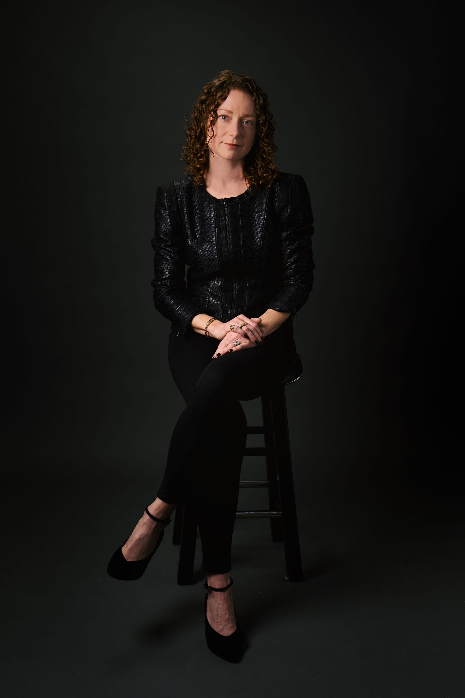Woman in black leather jacket and pants sitting on stool against dark background.