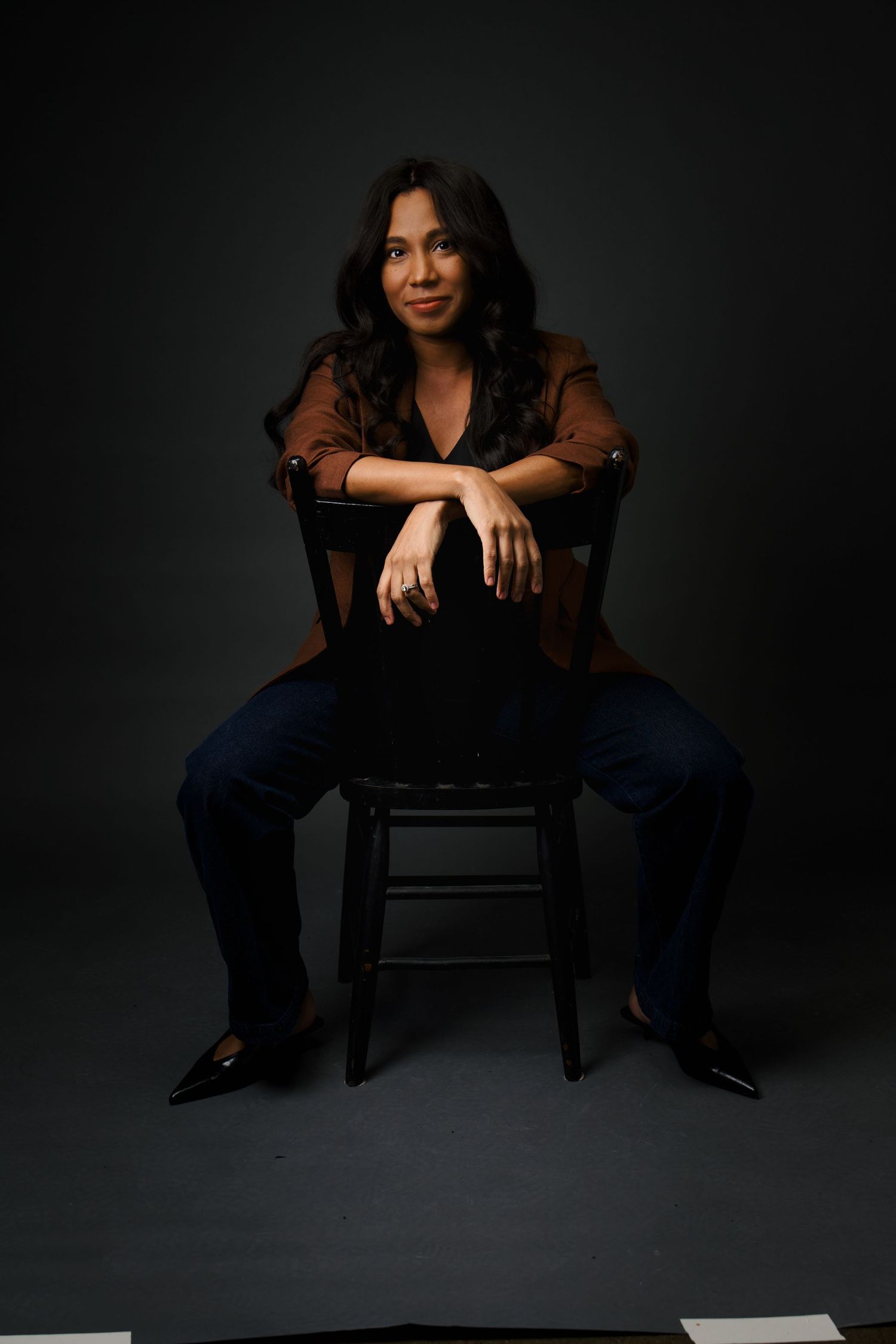 Woman in brown blouse and jeans sits on chair with arms crossed in dark studio.