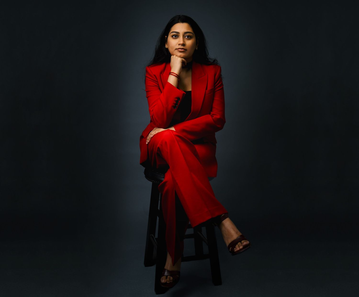 Woman in red suit seated on stool posing confidently against dark teal background.