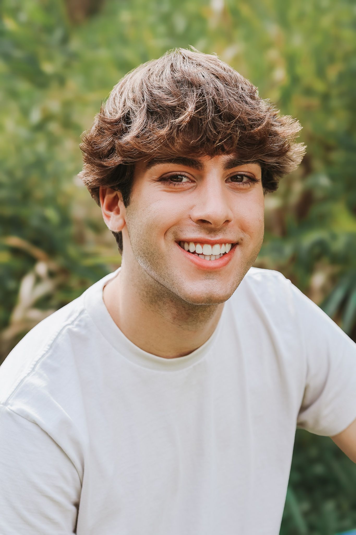 Cheerful young man with curly hair smiling in white t-shirt against green foliage background.