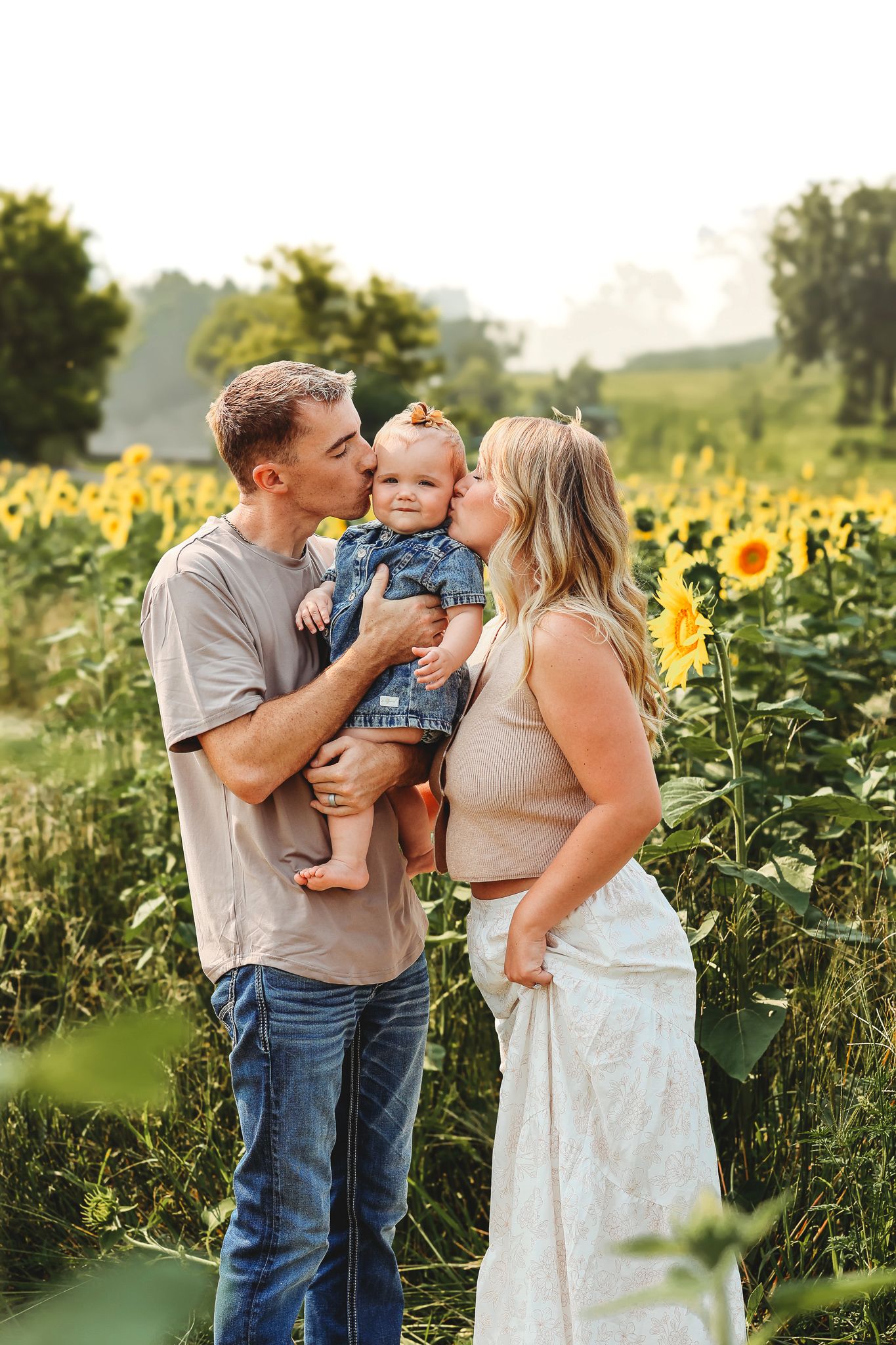 Parents kissing their baby on cheeks in a vibrant sunflower field on a warm summer day.