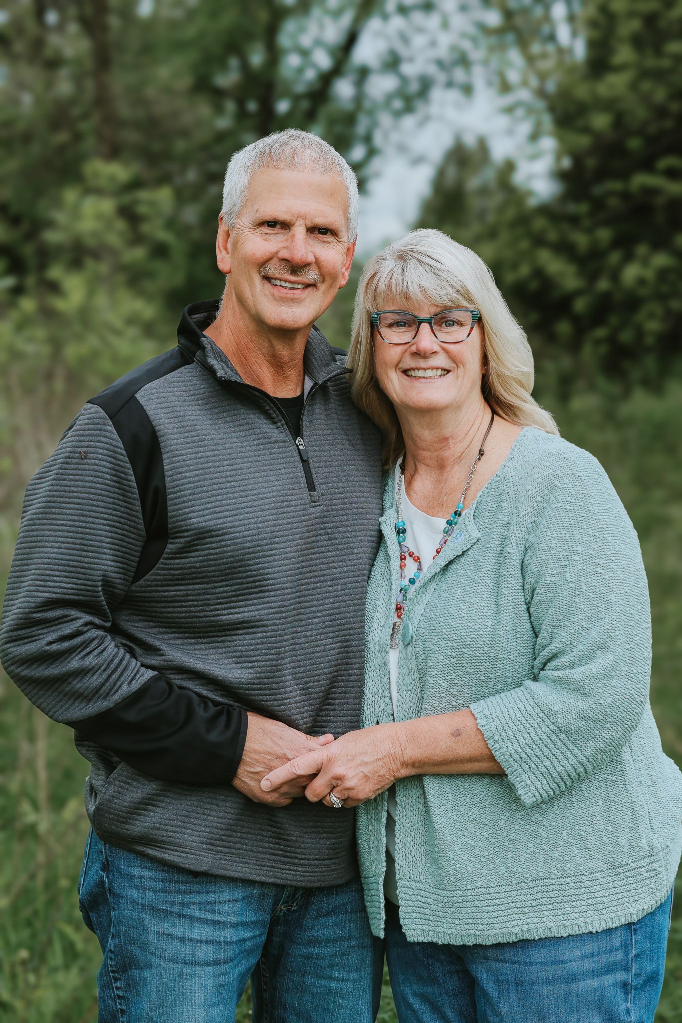A smiling older couple poses together outdoors surrounded by lush green trees.