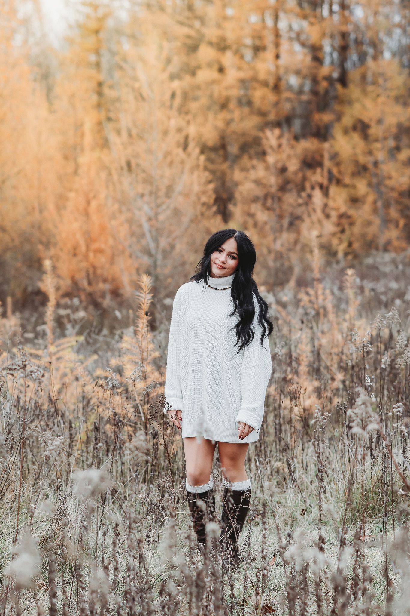Dark-haired young woman in white oversized sweater and tall boots standing in a frost-touched autumn field.