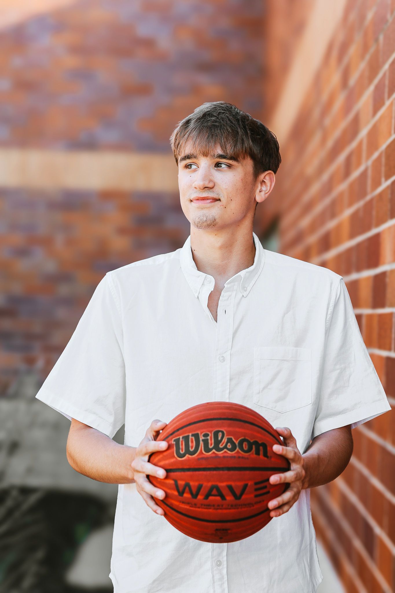 Young man in white button-up shirt holding a Wilson Wave basketball near a brick wall.