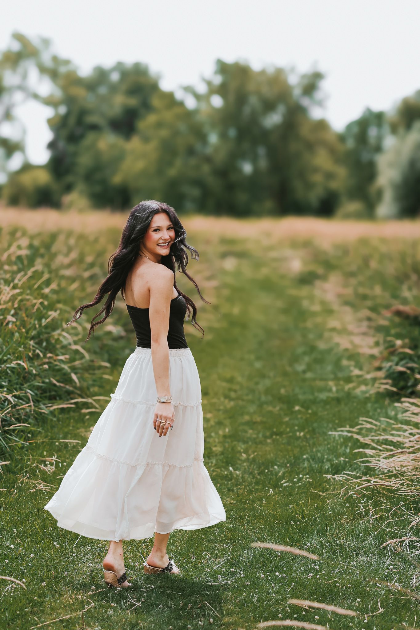 Young woman with long dark hair twirling in a white maxi skirt and black top in an open field at golden hour.