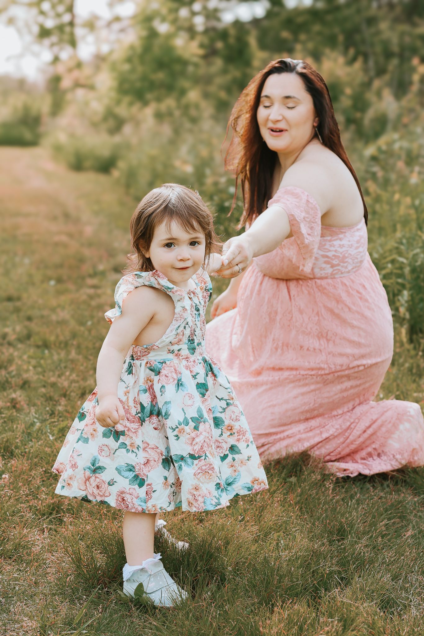 Pregnant woman in pink lace dress holding hands with toddler girl in floral dress in a sunny outdoor setting.