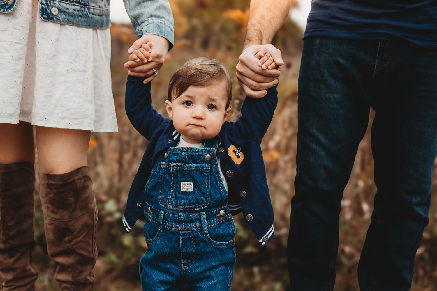 Baby in denim overalls holds hands with parents in an autumn outdoor setting.