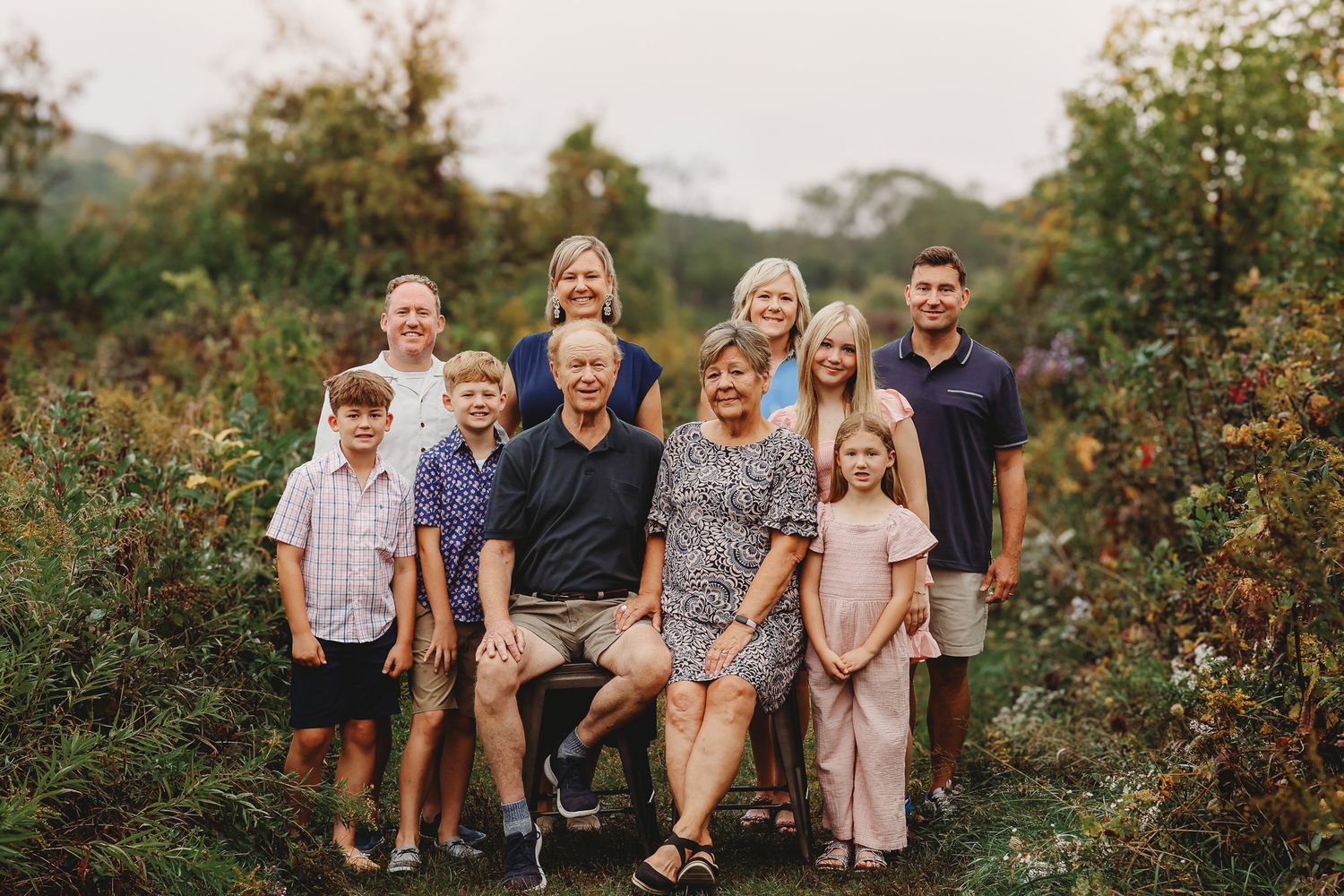 Large extended family poses together outdoors among lush green and golden foliage.