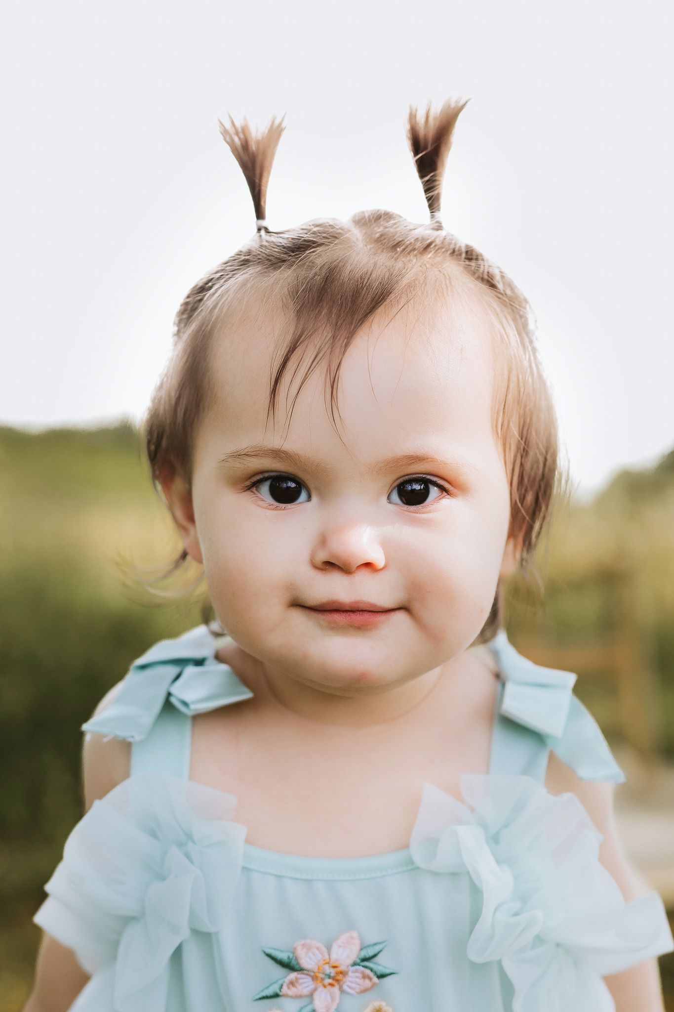 Close-up portrait of a baby girl with tiny pigtails wearing a light blue floral dress outdoors.