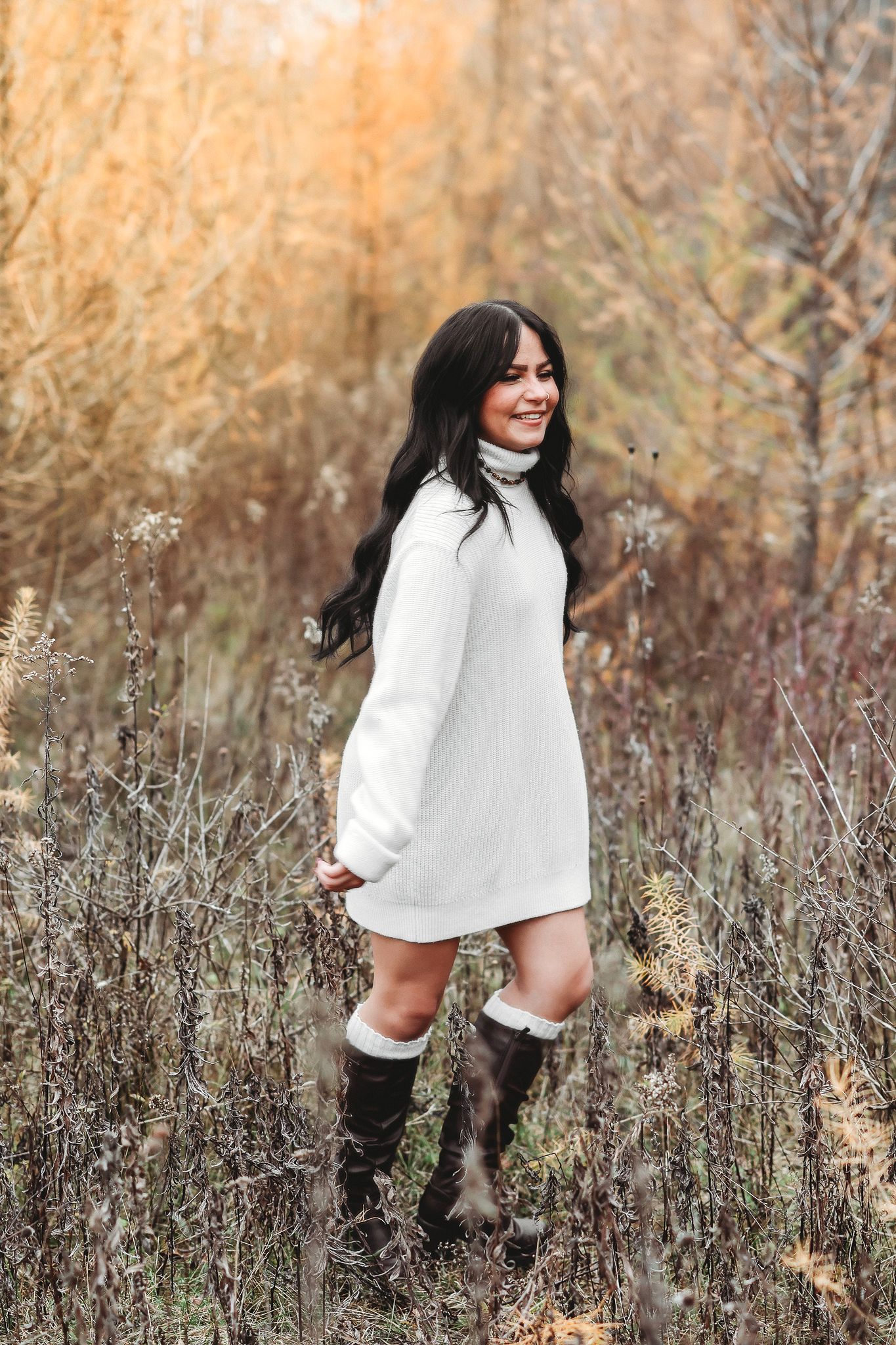 Smiling brunette woman in white sweater dress and tall brown boots standing in fall meadow.