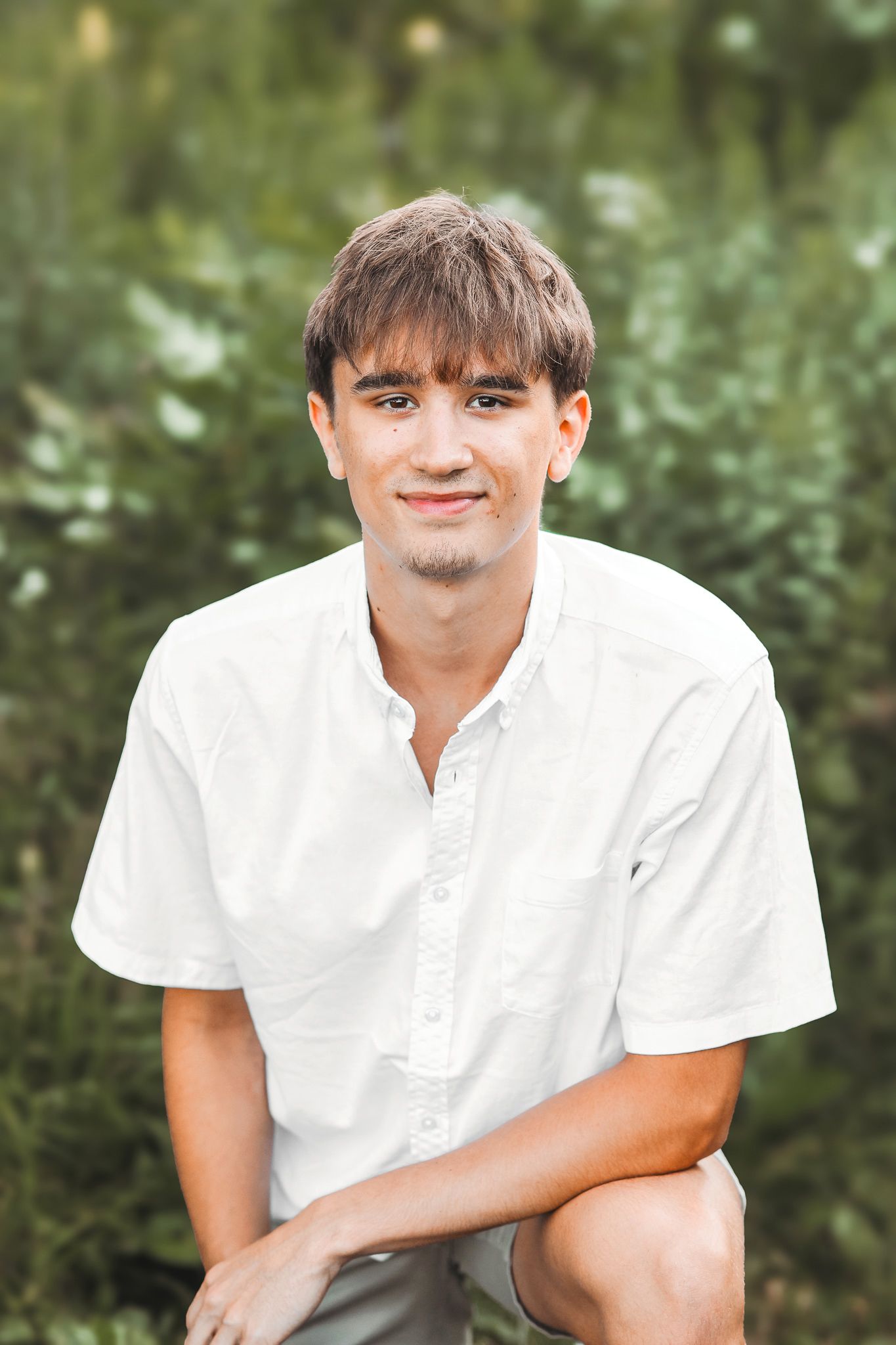 Smiling young man in white short-sleeve shirt posing outdoors with green foliage background.