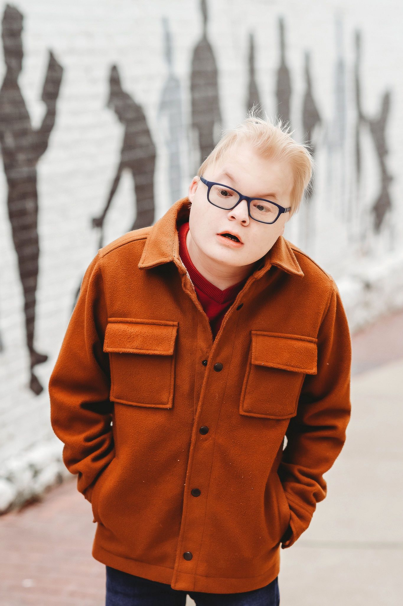 Blonde boy with Down syndrome wearing brown jacket and glasses standing near graffiti wall.