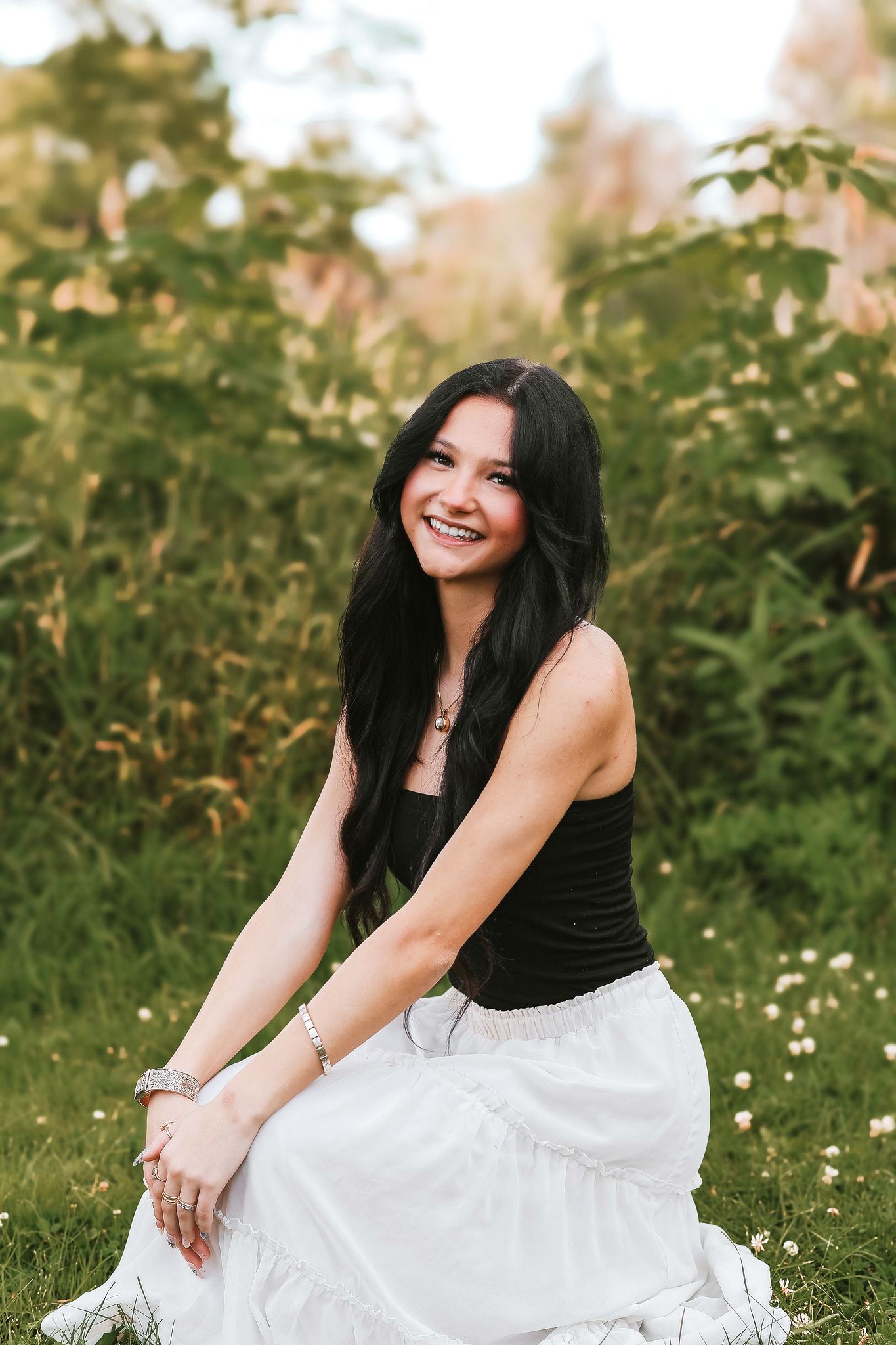 Happy young woman with long dark hair sitting on grass in a white skirt and black top surrounded by green foliage.