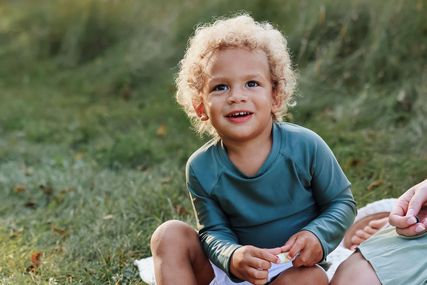 Curly-haired toddler in green shirt smiling outdoors on grass.