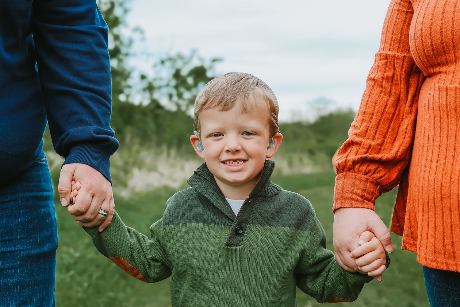 A smiling young boy in a green sweater holds hands with two adults in a field.