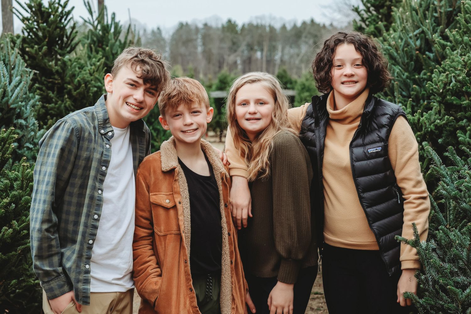 Four smiling children pose together at a Christmas tree farm in casual autumn outfits.