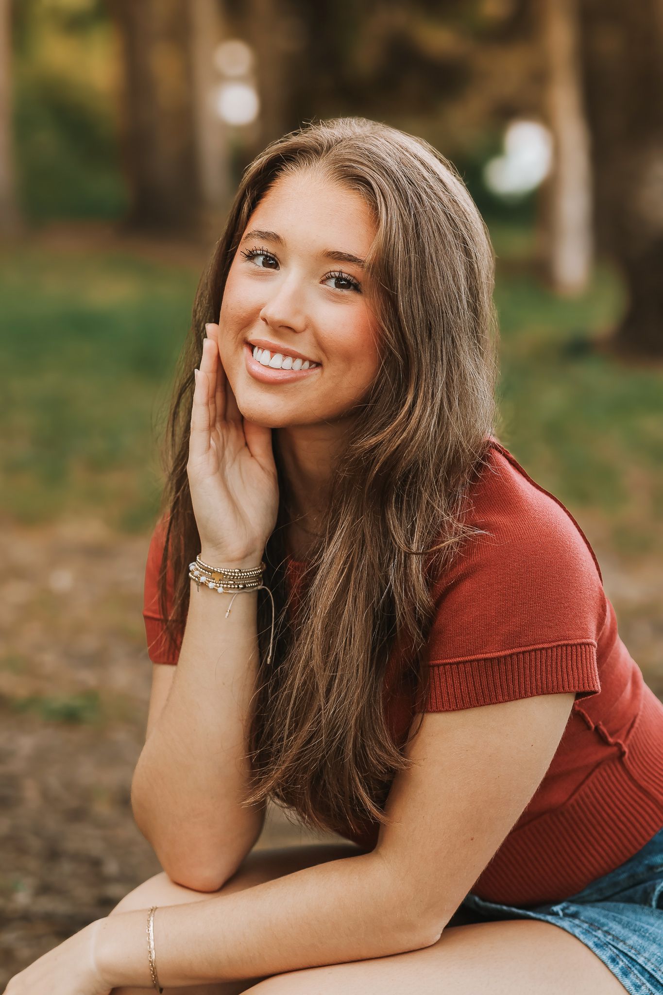 Smiling teenage girl in rust red top leaning on hand for outdoor senior portrait photo.