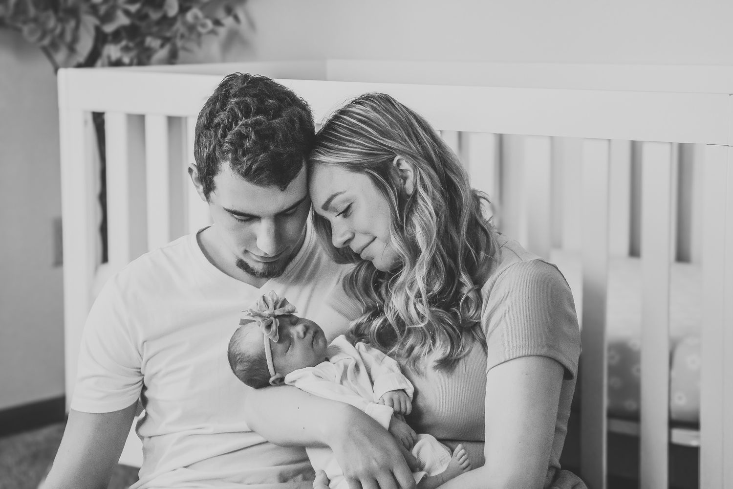 Black and white photo of loving parents cradling their newborn baby together near a white crib.