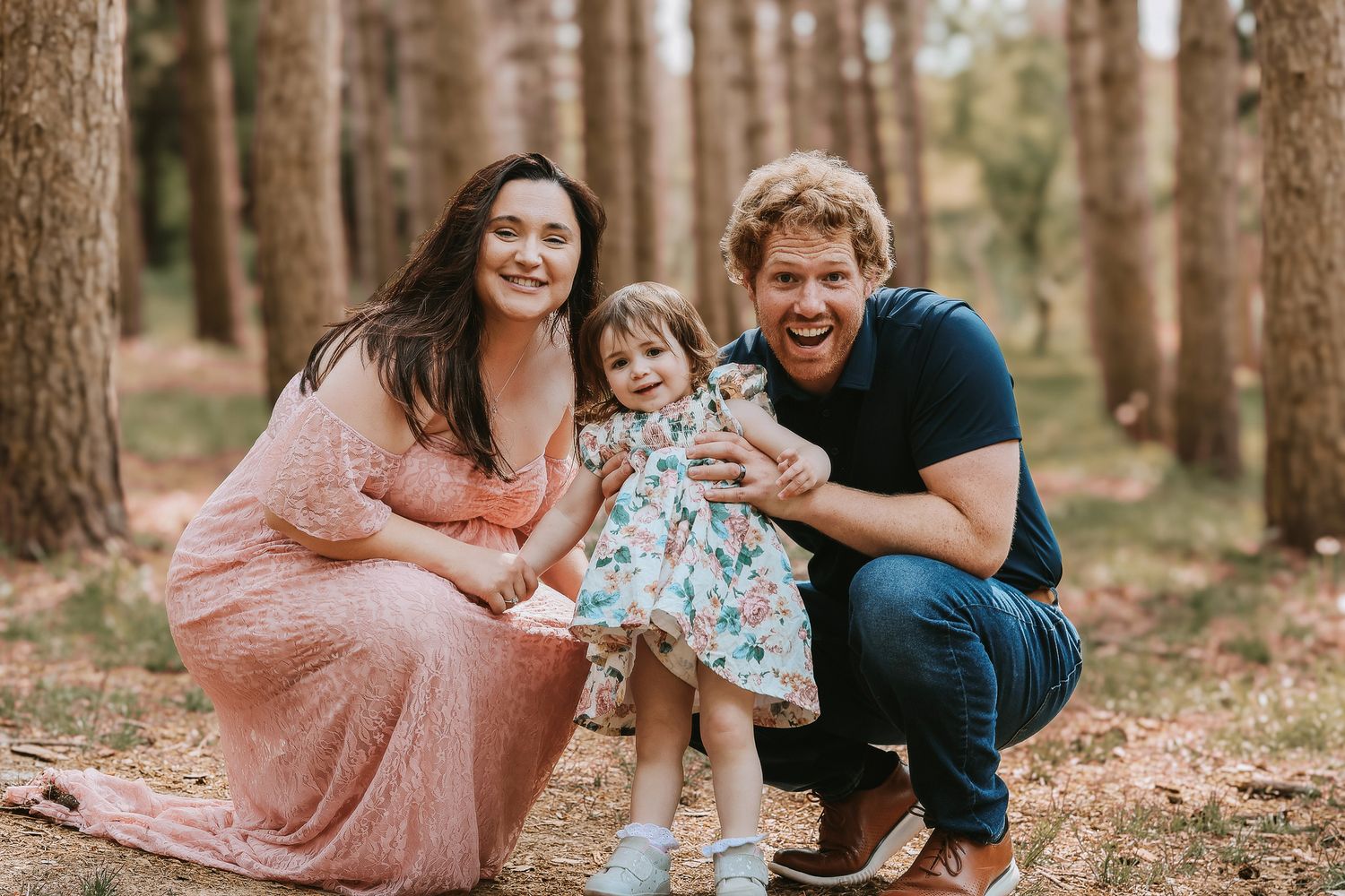 Happy family of three posing together in pine forest, mom in pink dress, dad and toddler daughter smiling.