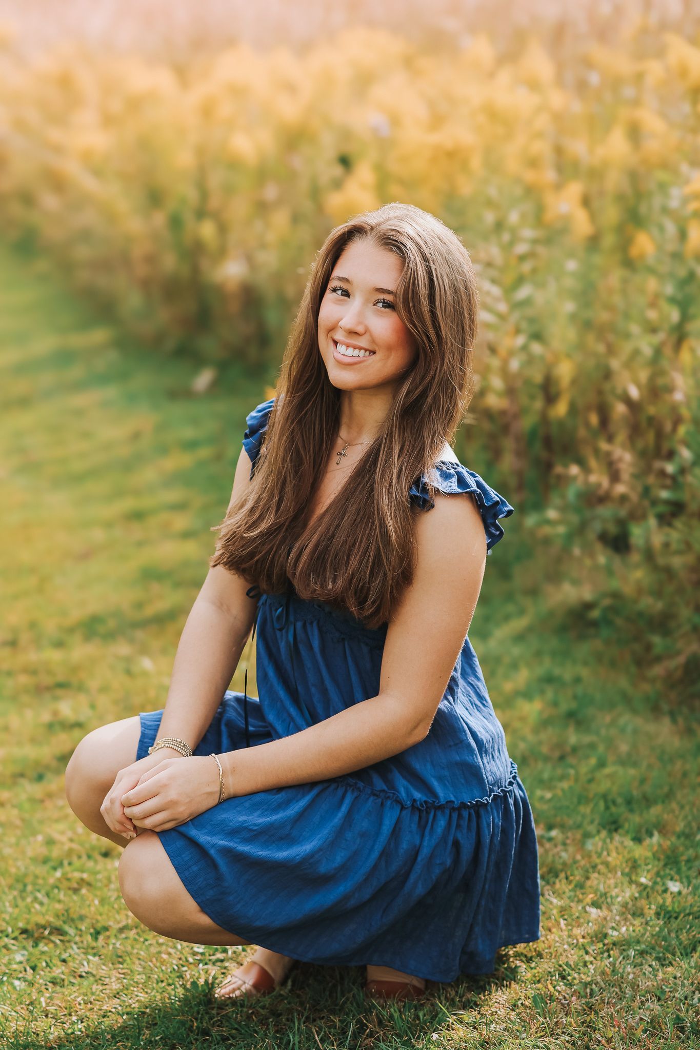 Smiling teenage girl in blue dress crouching on grass with yellow wildflowers behind her.
