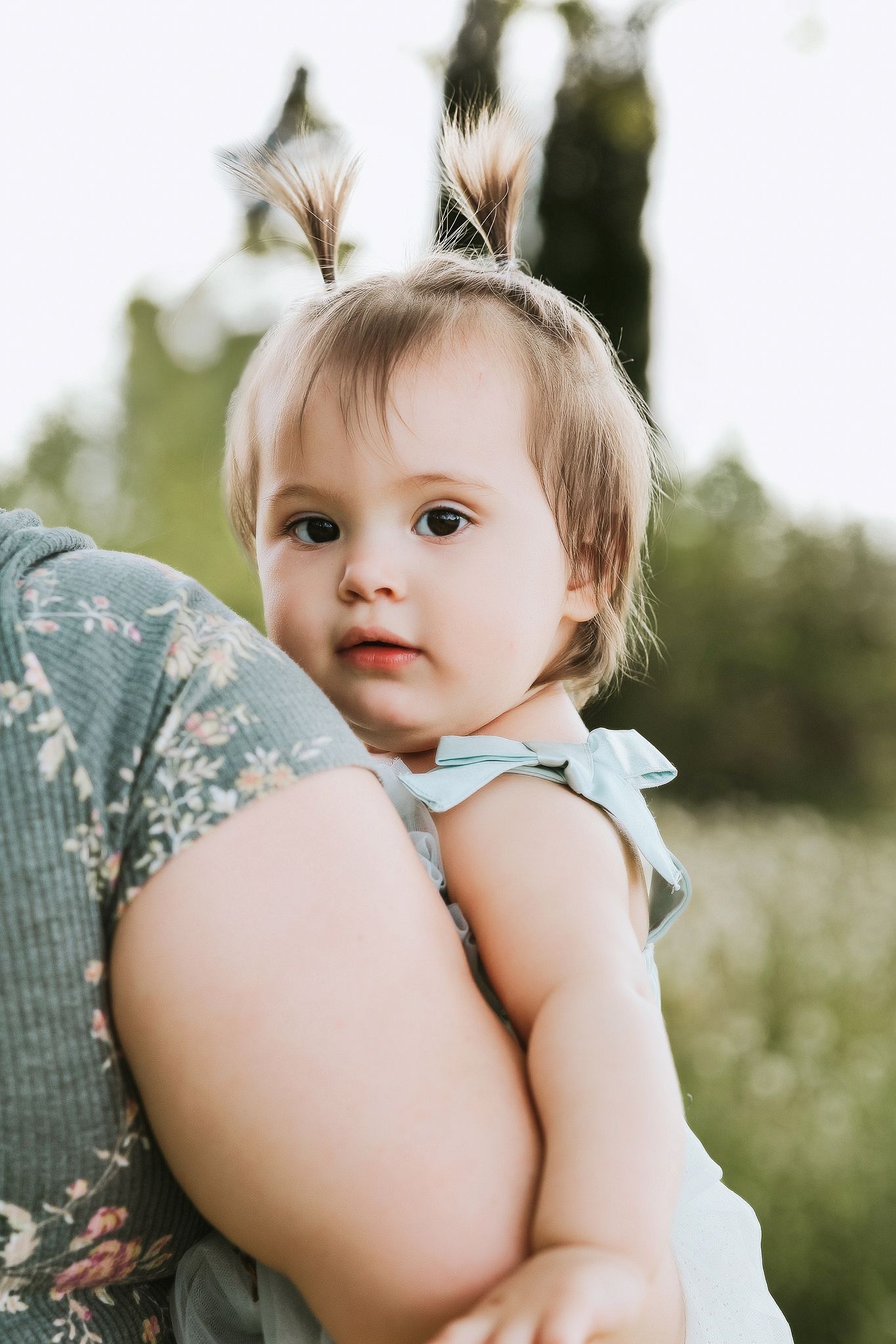 Sweet baby girl in light blue dress being held by an adult in a sunlit outdoor setting.