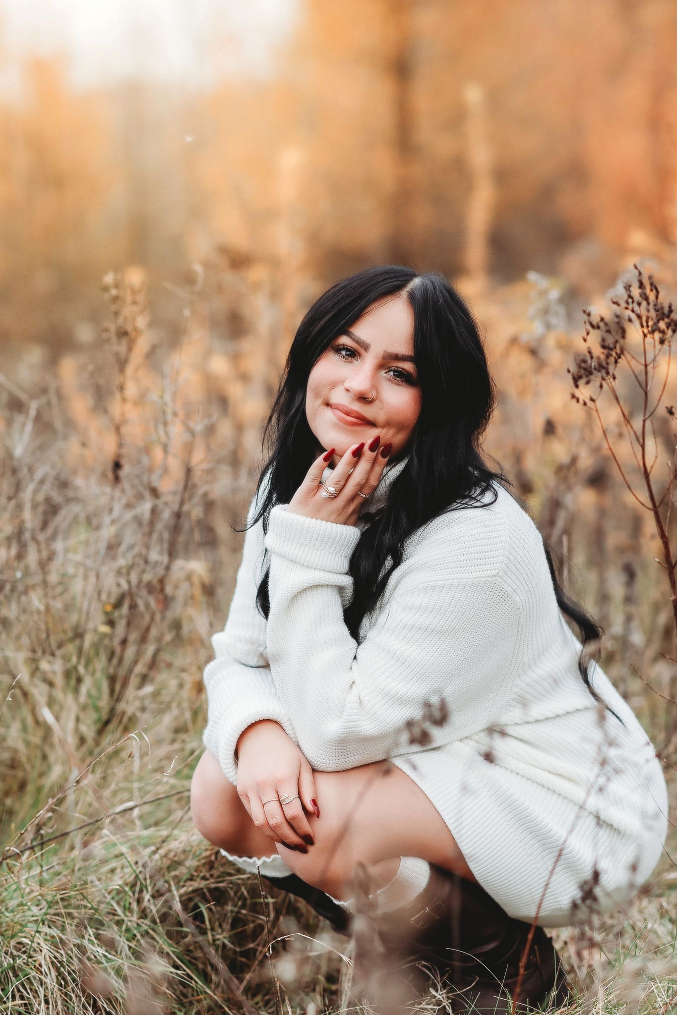 Smiling young woman with black hair in white sweater posing in dry autumn grass field.