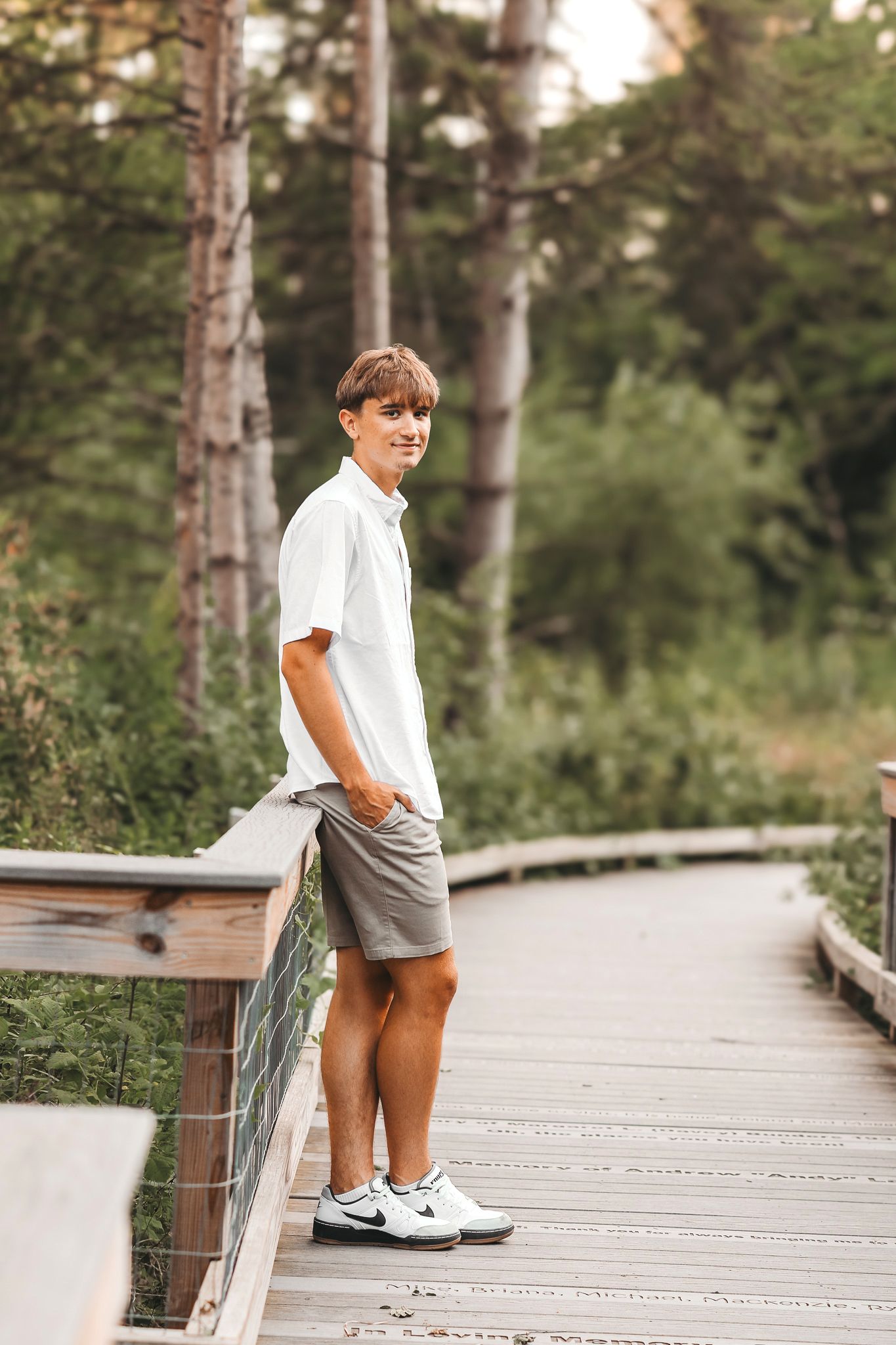Teen boy in white shirt and gray shorts standing on a wooden boardwalk surrounded by trees.