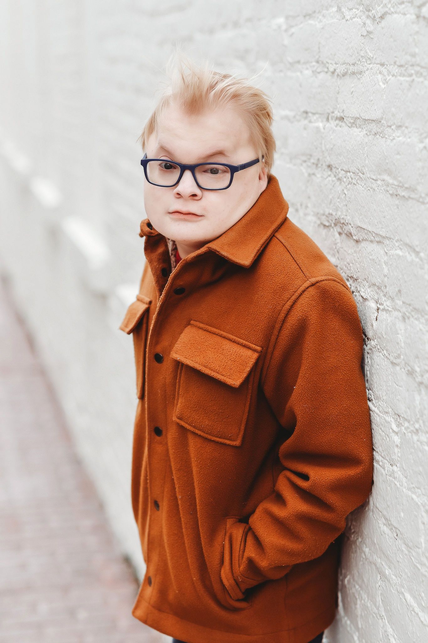 Blonde boy with Down syndrome in rust brown jacket leaning against white brick wall outdoors.